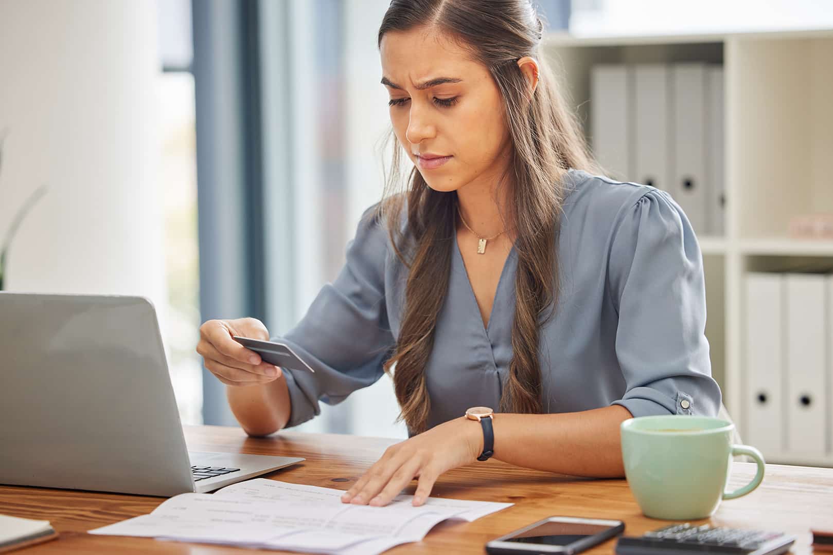 Fiatal ingatlanvagyon-értékelő és -közvetítő nő ül az irodában egy laptop előtt, és koncentráltan néz egy bankkártyát. ingatlanvagyon-értékelő és -közvetítő képzés a Pannon Kincstárban!