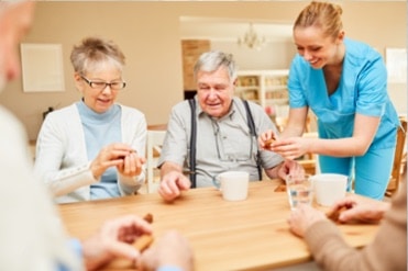 A demencia gondozó hölgy kék nővérruhában segíti az asztalnál ülő idős demens betegeket.
