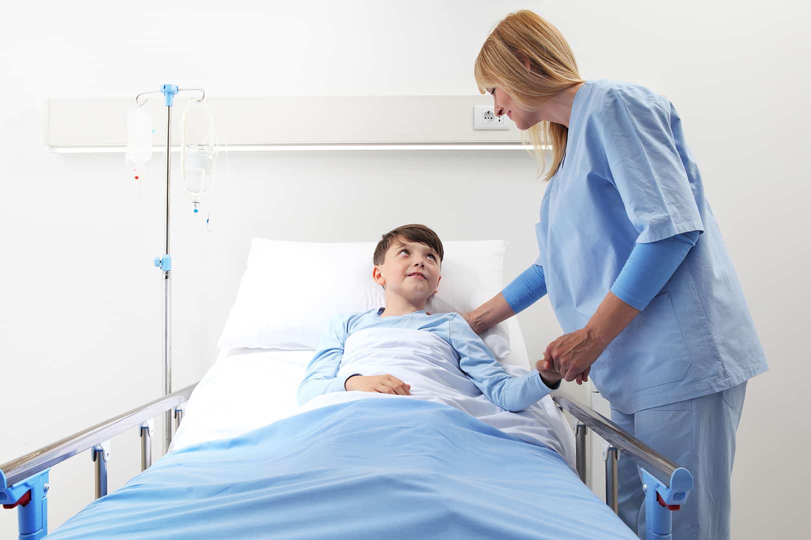 Gyermek intenzív szakápoló nő egy kisfiú betegágya mellett áll, fogja a kezét, és mosolyog. Gyermek intenzív szakápoló képzés a Pannon Kincstárban!
