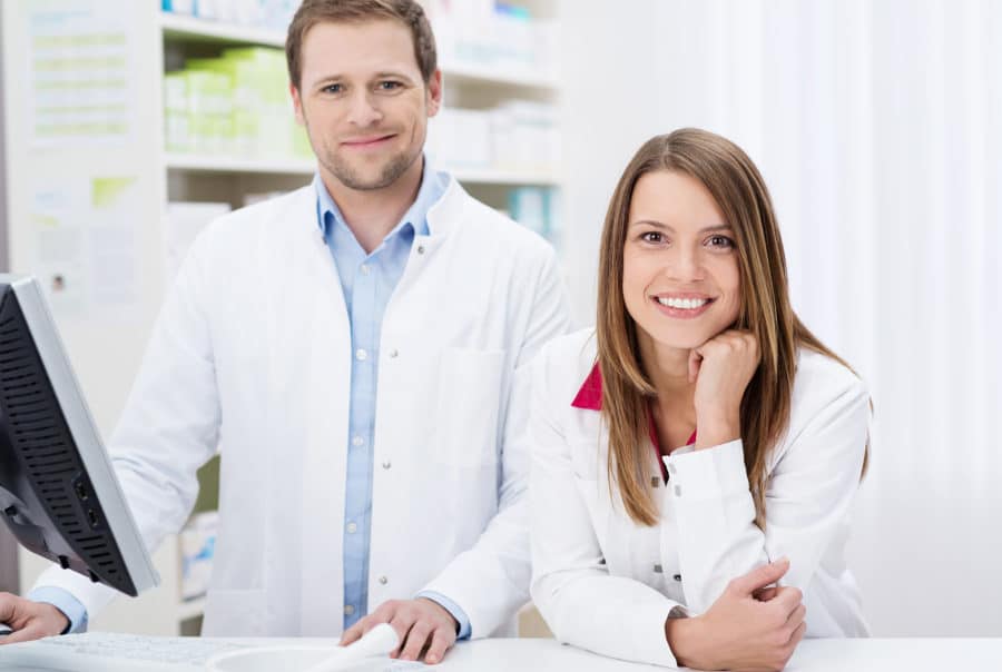 Egy férfi és egy nő gyógyszertári szakasszisztens áll a gyógyszertárban, fehér köpenyben a pénztárnál.