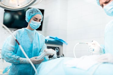 Aneszteziológiai szakasszisztens hölgy kék műtét közben viselt orvosi felszerelésben segíti az altatóorvos munkáját.