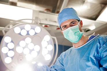 A műtéti szakasszisztens férfi, kék orvosi ruhában, maszkban áll egy nagy lámpa alatt.