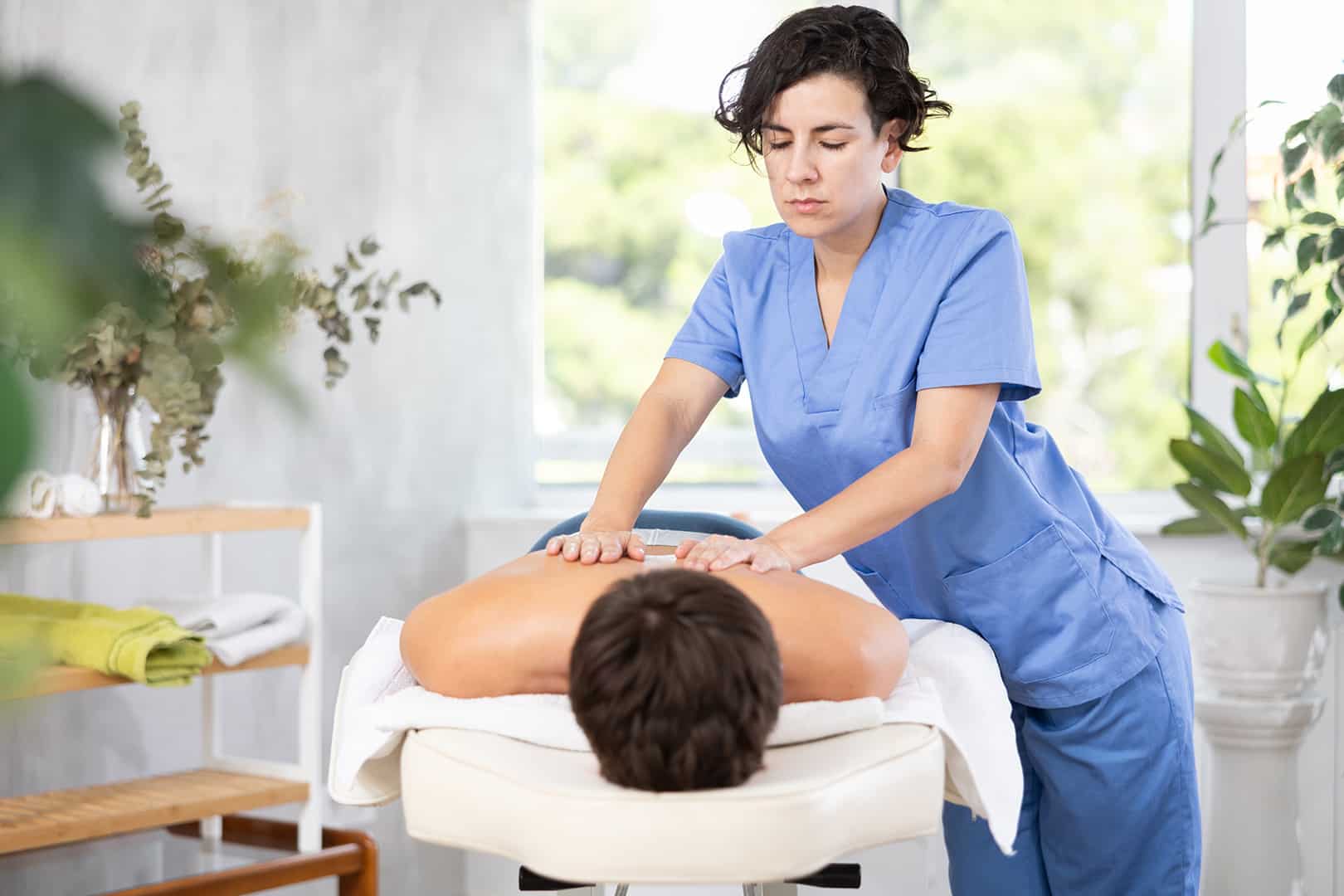 Gyógymasszőr nő orvosi ruhában masszírozza egy férfi hátát. Gyógymasszőr képzés a Pannon Kincstárban!