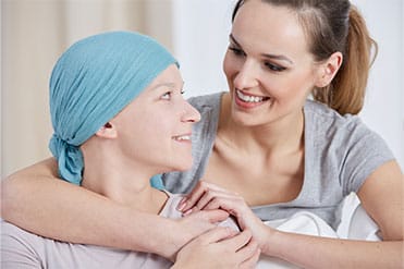 Barna hajú hospice szakápoló, palliatív gondozó ölel egy kék fejkendős lányt egy világos szobában.