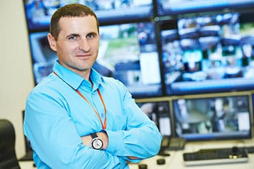 Egy biztonságszervező férfi kék ingben, ölbe tett kézzel áll több monitor előtt.