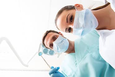 Két klinikai fogászati higiénikus kék orvosi ruhában, gumikesztyűben és maszkban figyelik felülről páciensüket.