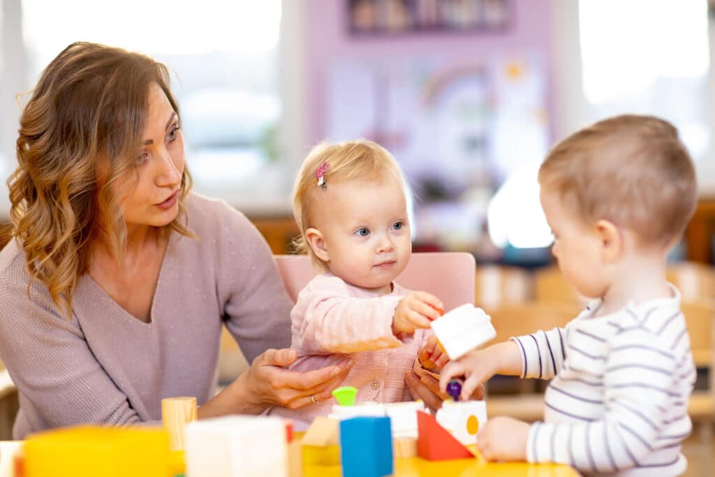 Szociális munkás nő egy asztalnál ülve két kisgyermekkel játszik építőkockákkal.