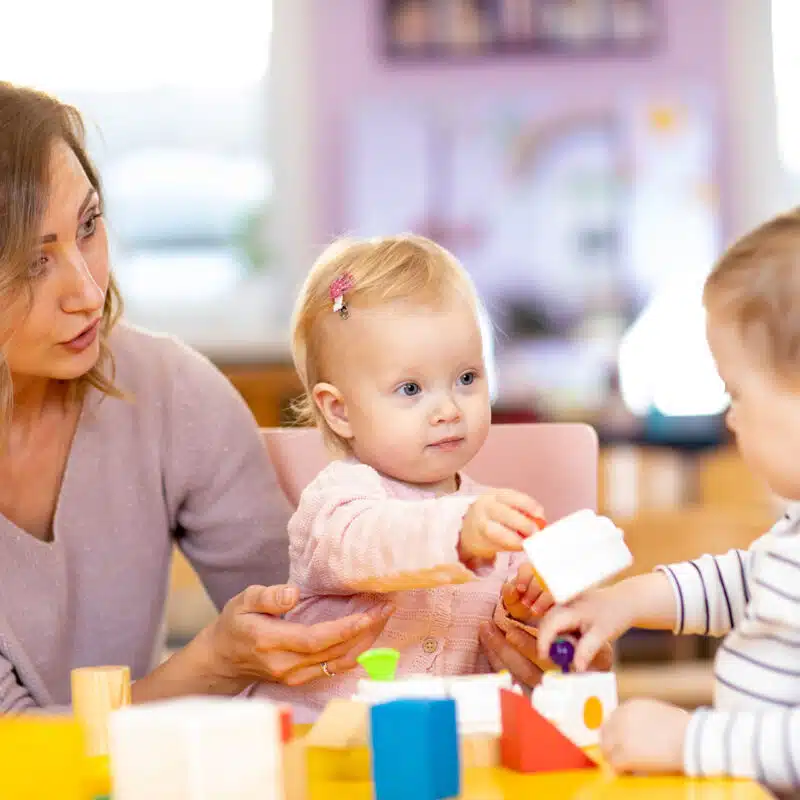 Szociális munkás nő egy asztalnál ülve két kisgyermekkel játszik építőkockákkal.