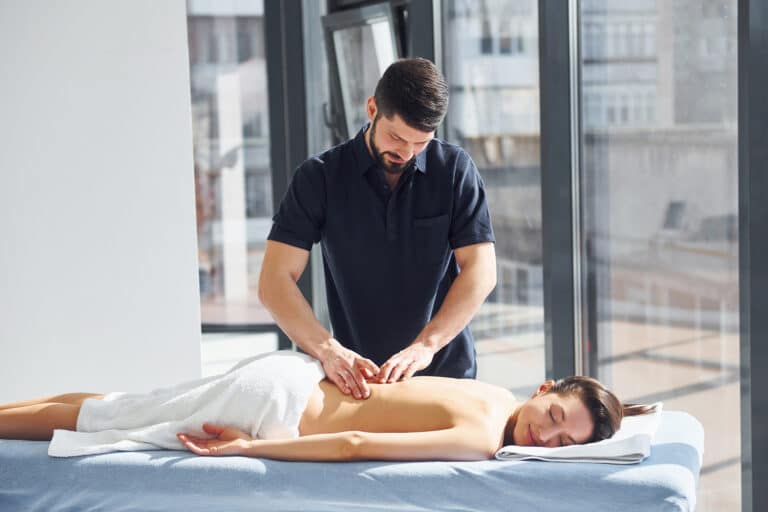 Gyógymasszőr férfi egy nőt masszíroz letisztult környezetben. Gyógymasszőr képzés, Pannon Kincstár