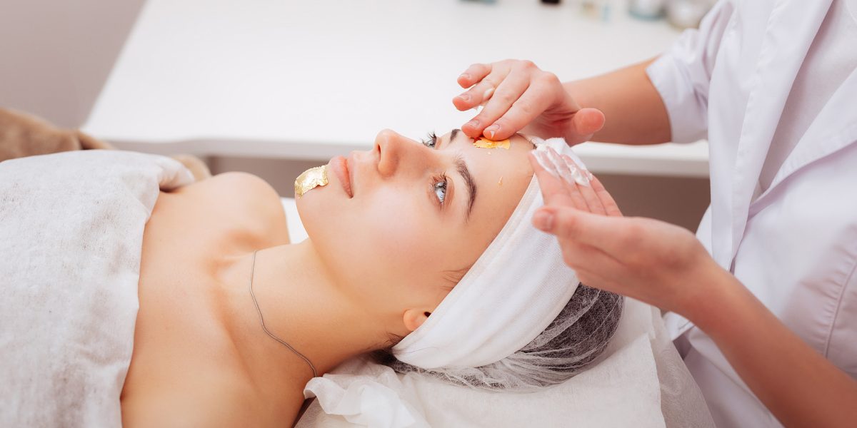 Full of minerals. Professional female cosmetologist holding golden mask sheets while applying them on her clients face