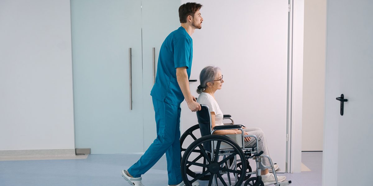 Young health worker accompanies an elderly patient in a wheelchair, the room is bright and clean