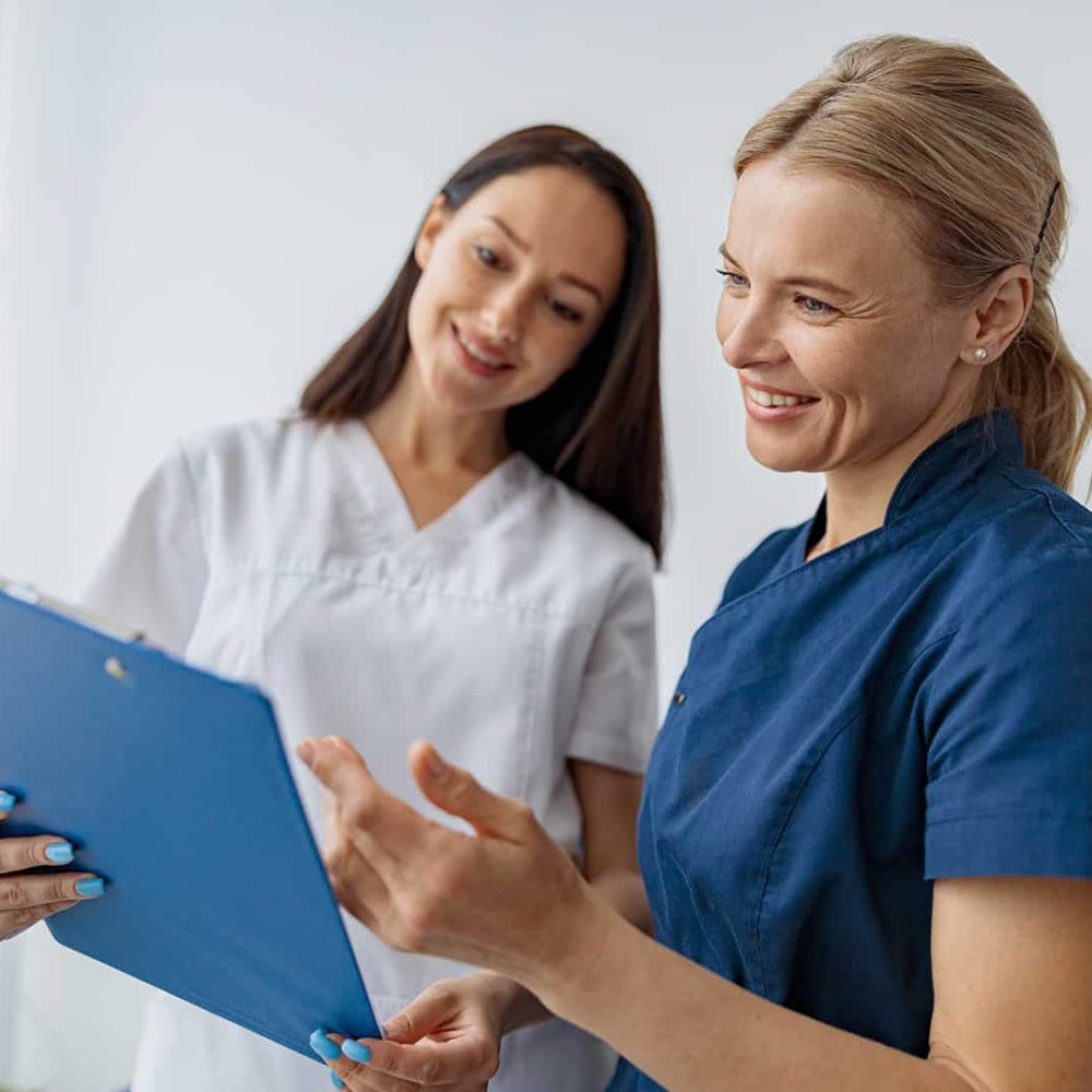 Egy kék egyenruhás általános ápoló és egy fehér ruhás doktornő mosolyogva néznek egy dokumentumot. Általános ápoló képzés a Pannon Kincstárban!