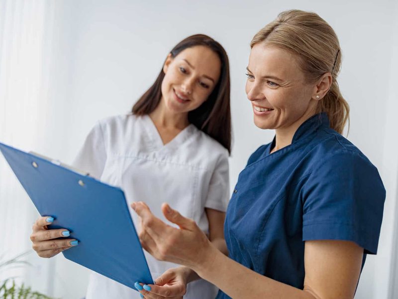 Kék egyenruhás általános ápoló és egy fehér ruhás doktornő mosolyogva néznek egy dokumentumot. Általános ápoló képzés a Pannon Kincstárban!