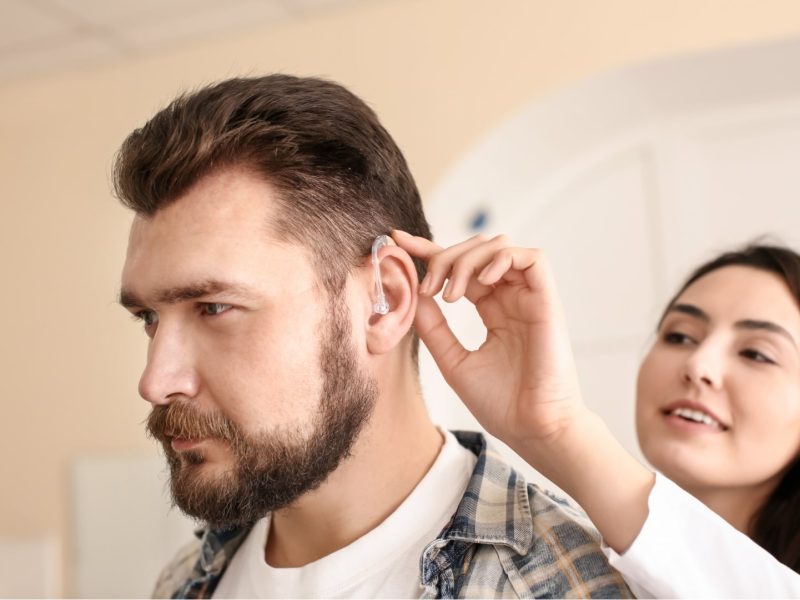 Mosolygós audiológiai asszisztens egy fiatal férfi hallásjavító készülékét igazítja