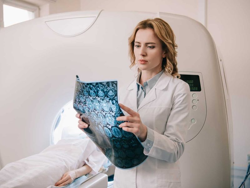 CT/MR szakasszisztens képzés Egy fiatal asszisztens nő fehér ruhában MRI felvételeket vizsgál, a háttérben egy páciens fekszik az MRI gépben.