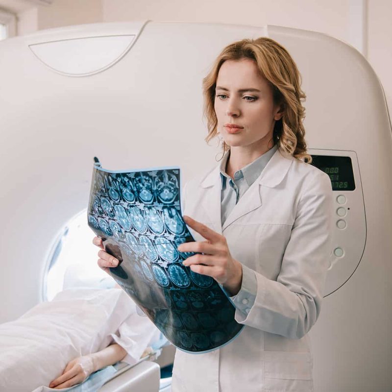 Fiatal asszisztens nő fehér ruhában MRI felvételeket vizsgál, a háttérben egy páciens fekszik az MRI gépben. CT/MR szakasszisztens képzés a Pannon Kincstárban!