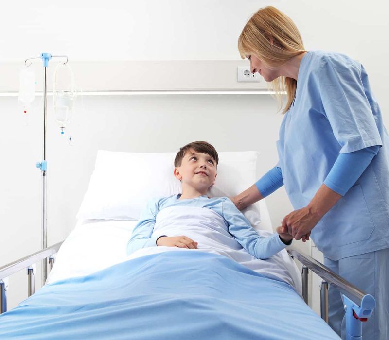 Gyermek intenzív szakápoló képzés, Pannon Kincstár Gyermek intenzív szakápoló nő egy kisfiú betegágya mellett áll, fogja a kezét, és mosolyog. Gyermek intenzív szakápoló képzés a Pannon Kincstárban!
