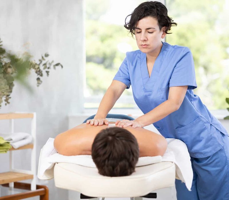 Gyógymasszőr képzés, Pannon Kincstár Gyógymasszőr nő orvosi ruhában masszírozza egy férfi hátát. Gyógymasszőr képzés a Pannon Kincstárban!