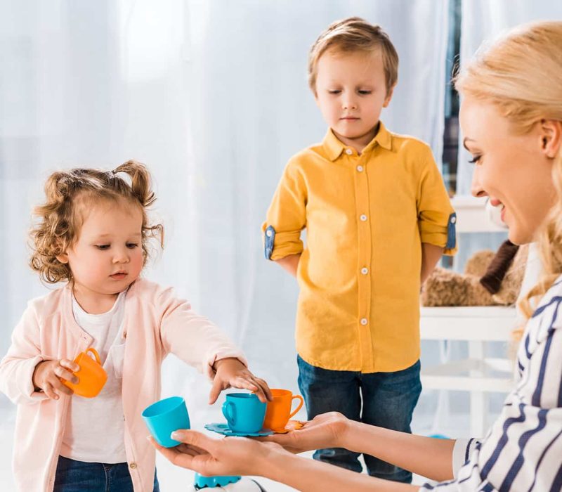 Egy mosolygós kisgyermekgondozó nő játszik egy kislánnyal és egy kisfiúval az óvodában.