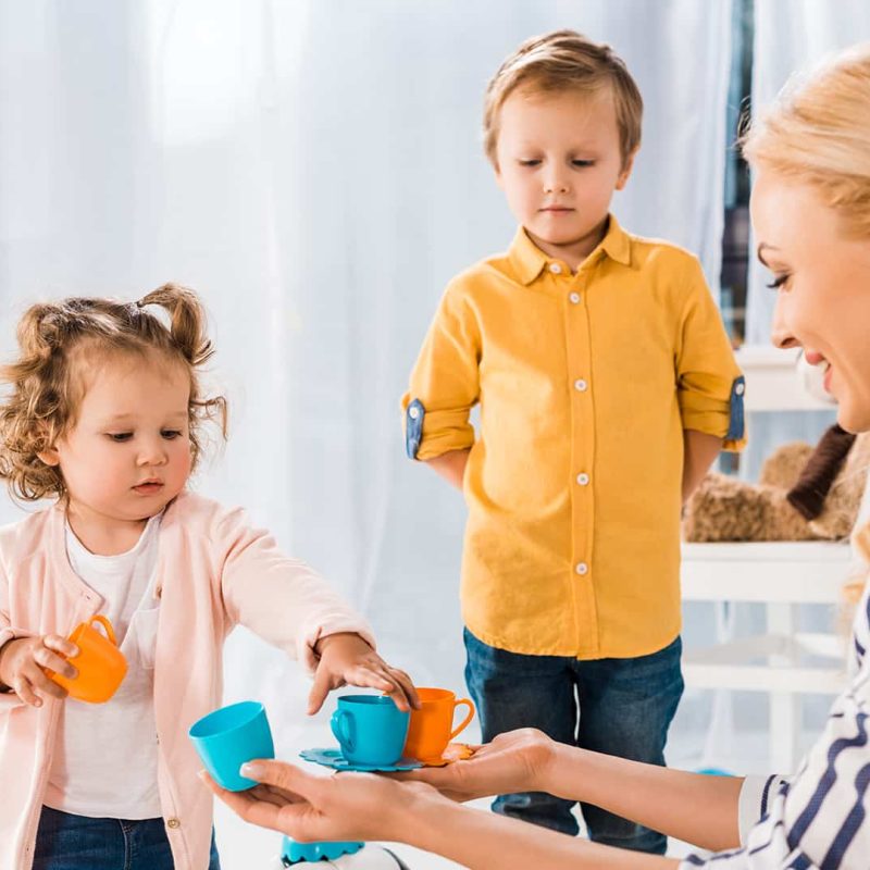Mosolygó kisgyermekgondozó nő játszik egy kislánnyal és egy kisfiúval az óvodában. Kisgyermekgondozó,-nevelő képzés a Pannon Kincstárban!