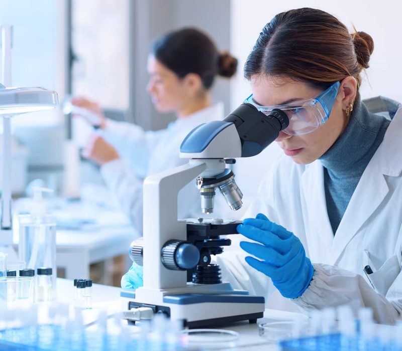 Egy fehér köpenybe és védőszemüvegbe öltözött szövettani asszisztens nő mikroszkópot használ egy laboratóriumban. Szövettani asszisztens képzés a Pannon Kincstárban!