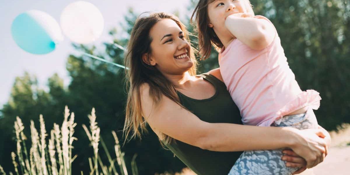Fiatal lány ül egy mezőn, kezében egy kislány van és két léggömb. Pannon Kincstár
