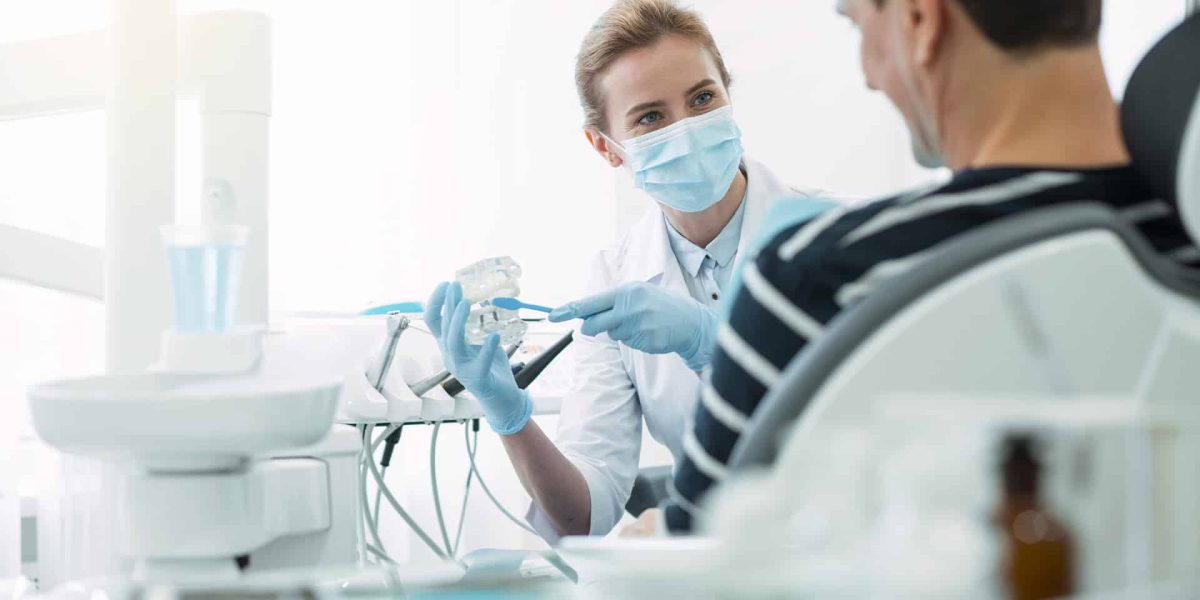 Klinikai fogászati higiénikus nő egészségügyi ruházatban, maszkban egy fogászati demonstrációs modellt tart a kezében és egy páciensnek mutatja. Klinikai fogászati higiénikus képzés a Pannon Kincstárban, szakmai cikk